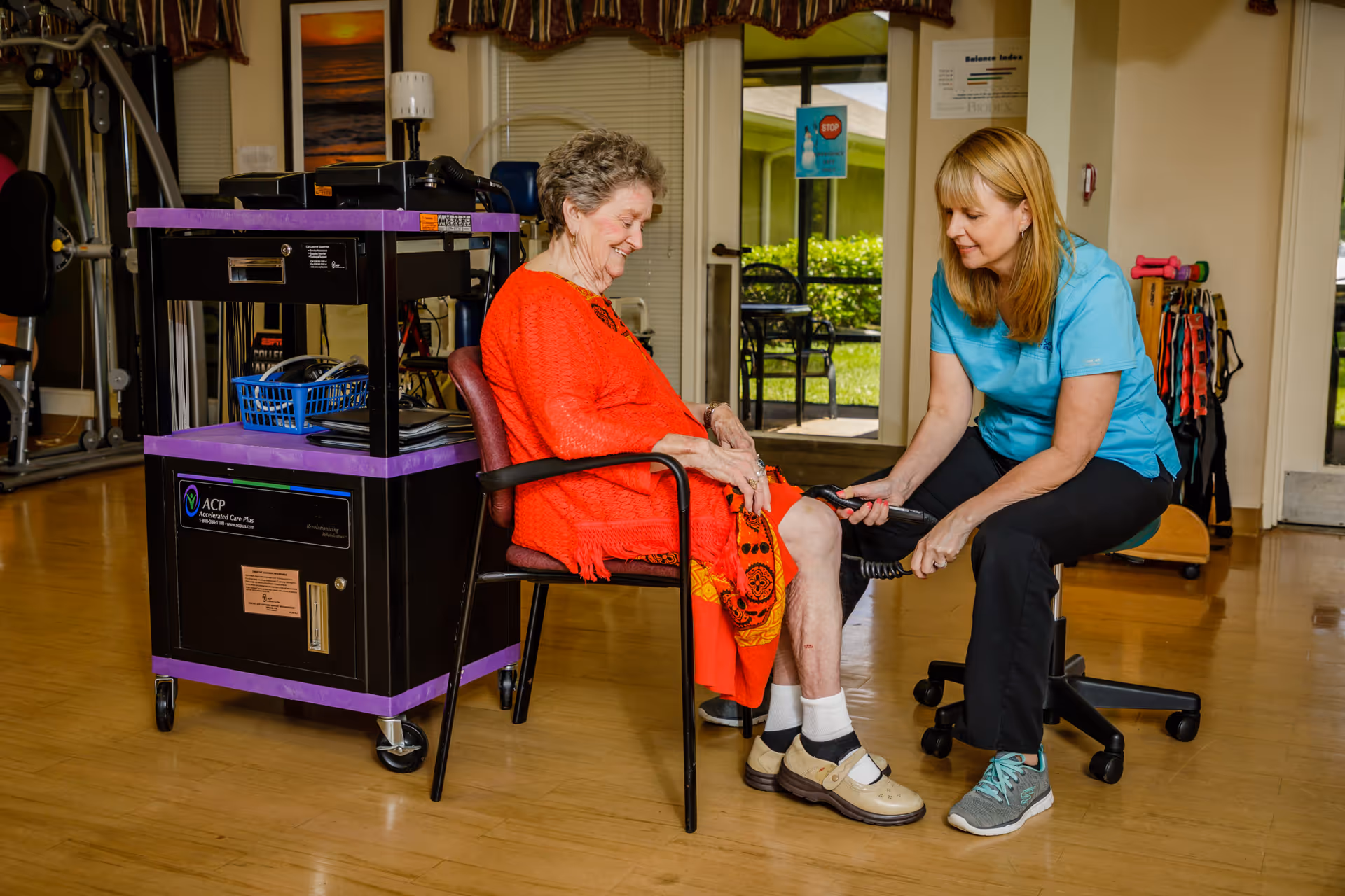 An elderly woman in a red dress sits on a chair while a healthcare worker in a blue shirt kneels in front of her, holding a device to her leg in a therapy or rehabilitation room with exercise equipment and a door leading outside.