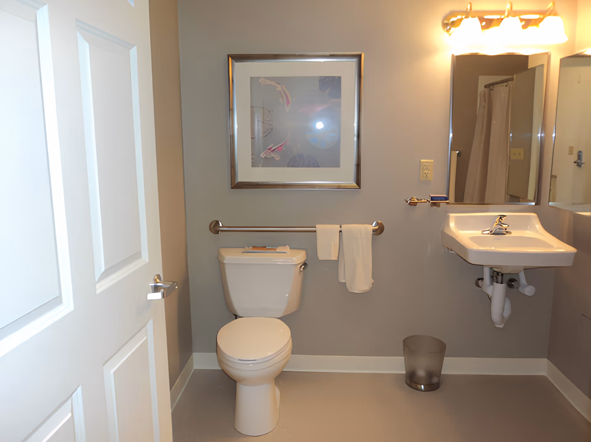 Small accessible bathroom with a toilet, wall-mounted sink, mirror, towel bar, and framed artwork on the wall.
