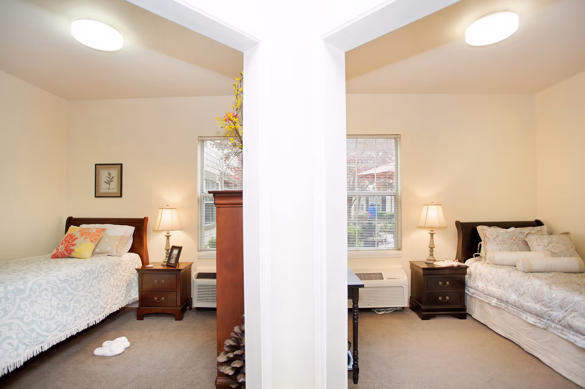 Two adjacent bedrooms separated by a white wall, each with a single bed, nightstand with a lamp, window with blinds, and carpeted floor. The left bedroom has a bed with a white and light blue patterned bedspread and colorful pillows, while the right bedroom has a bed with a beige patterned bedspread and multiple pillows.