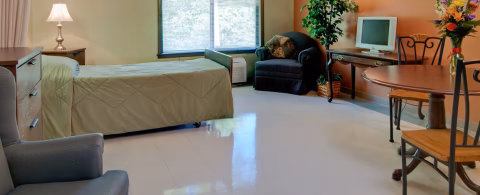 A nursing facility room with a single bed covered in a green quilt, a wooden nightstand with a lamp, a cushioned armchair, a wooden table with four chairs, a small TV on a wooden desk, and a large window letting in natural light. There are plants and a flower arrangement adding decoration to the room.