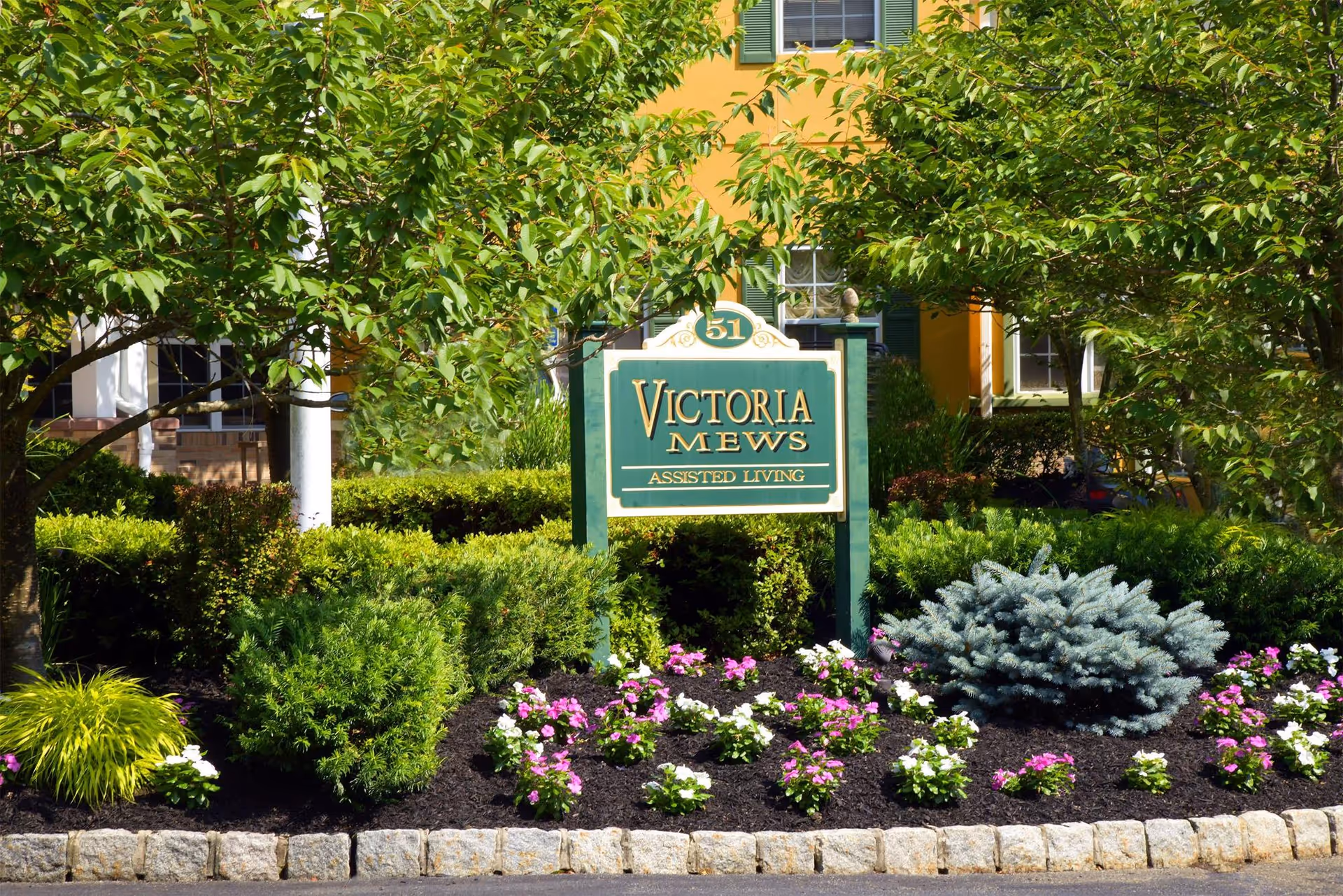 Landscaped front entrance sign reading "Victoria Mews Assisted Living" surrounded by shrubs, trees, and flowers.