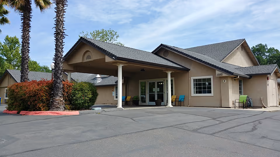 Exterior view of a single-story assisted living facility building with a covered entrance supported by white columns. There are several colorful chairs placed near the entrance and windows. Palm trees and bushes are visible near the building, and the sky is partly cloudy.