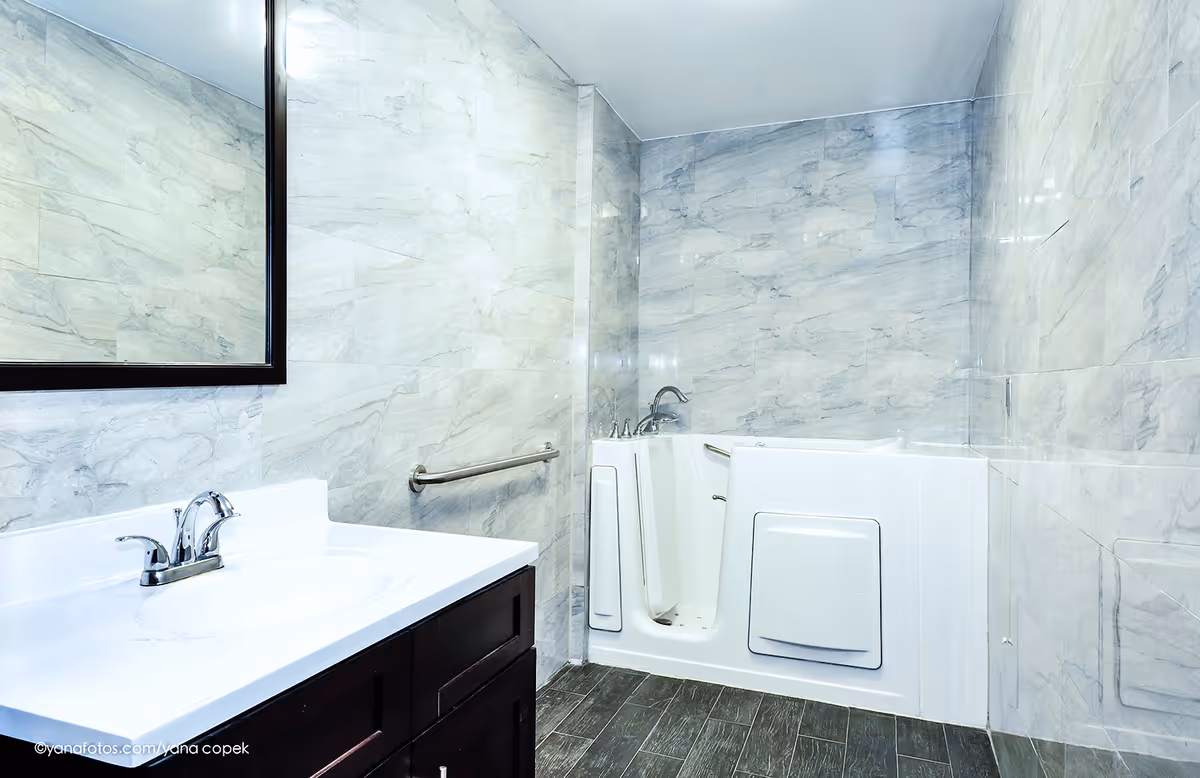 Bright accessible bathroom with a walk-in tub, vanity with sink and mirror, and tiled walls and floor.