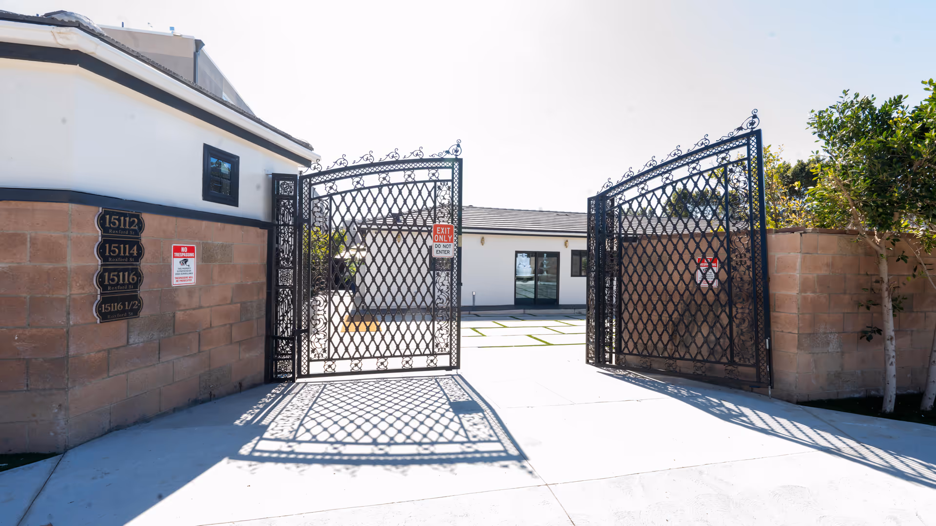 Open decorative black iron gates leading to a courtyard with a white building in the background. The gates cast intricate shadows on the concrete driveway. There are brick walls on either side of the gates with address plaques and no trespassing signs.