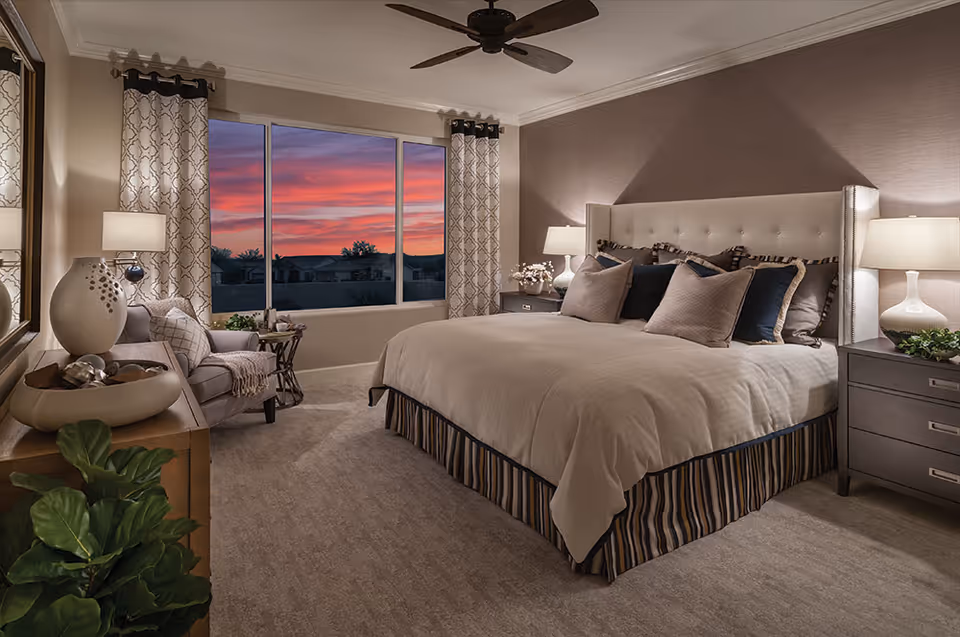 A well-decorated bedroom with a large bed featuring multiple pillows, two bedside tables with lamps, a comfortable armchair with a throw blanket, a wooden dresser with decorative items, and a large window showing a colorful sunset sky.