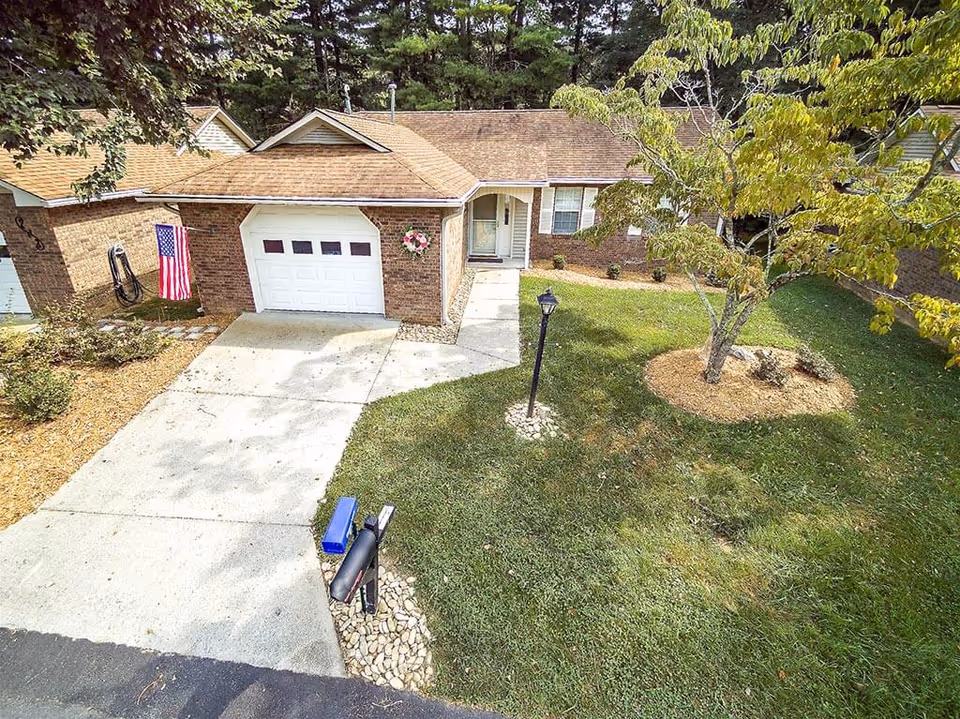 Front exterior of a single-story brick house with a driveway and single-car garage, an American flag, mailbox, lamp post, and a small grassy yard with a tree.