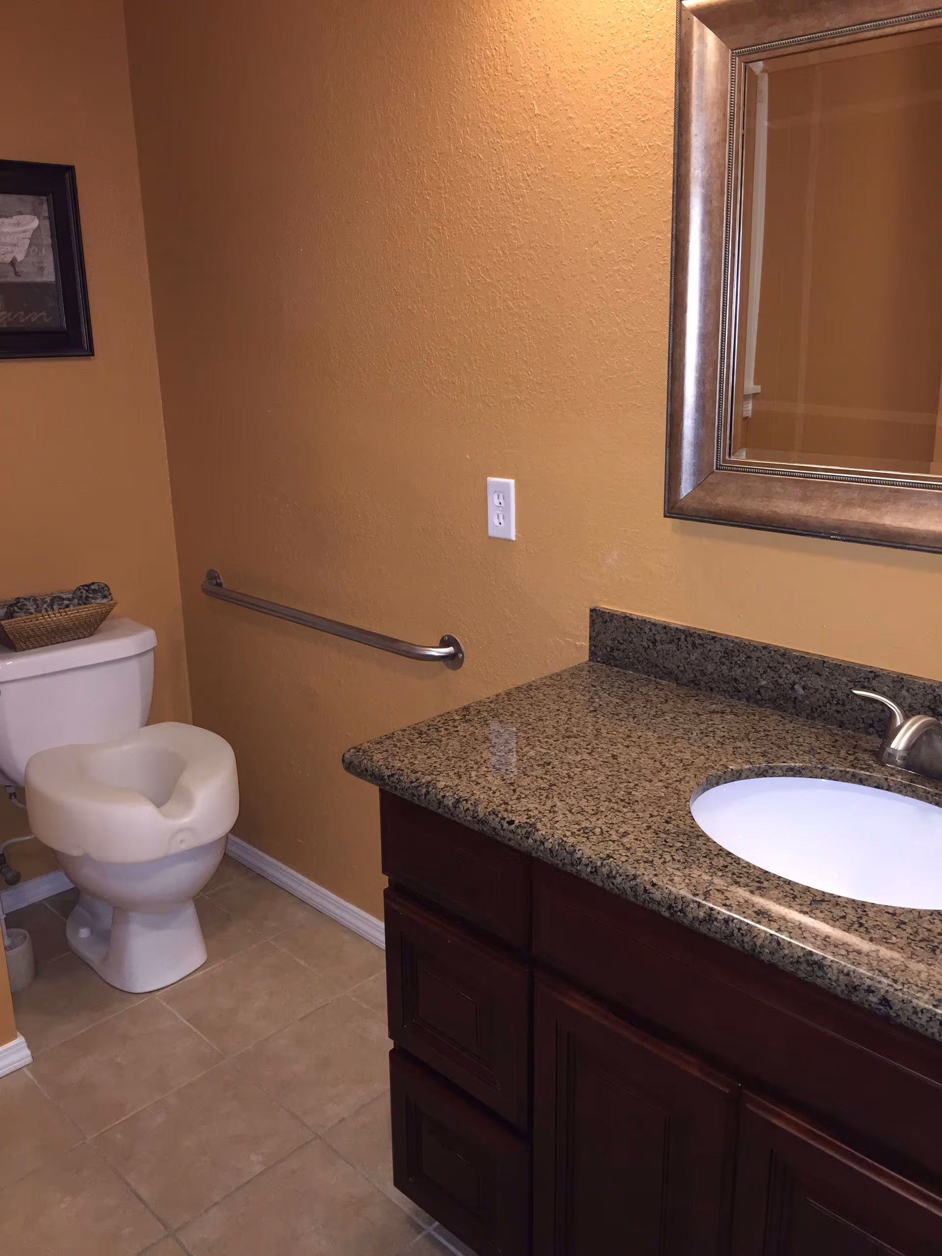 A bathroom with beige walls and tiled floor featuring a toilet with a padded seat, a granite countertop with an under-mount sink, a silver faucet, a large framed mirror above the sink, and a metal grab bar mounted on the wall next to the toilet.