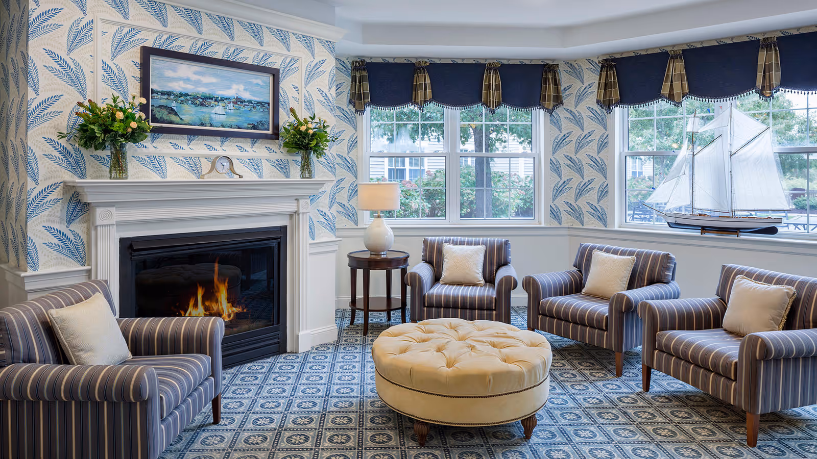A cozy living room with a lit fireplace, four striped armchairs each with a white cushion, a round tufted beige ottoman in the center, a side table with a lamp, and large windows with dark valances. The walls have blue leaf-patterned wallpaper and a framed painting above the fireplace. A model sailboat is displayed on the windowsill.