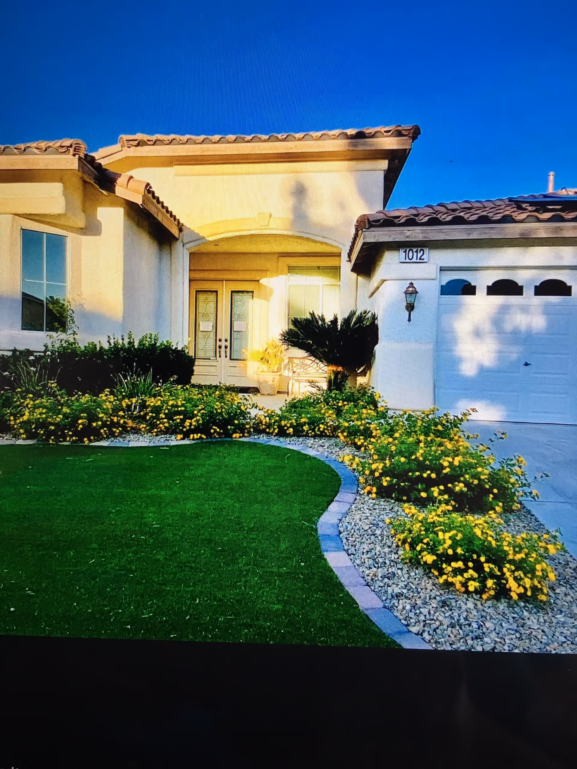 Front exterior of a single-story stucco house with a tiled roof, double front doors, garage, manicured lawn and yellow flowering shrubs.