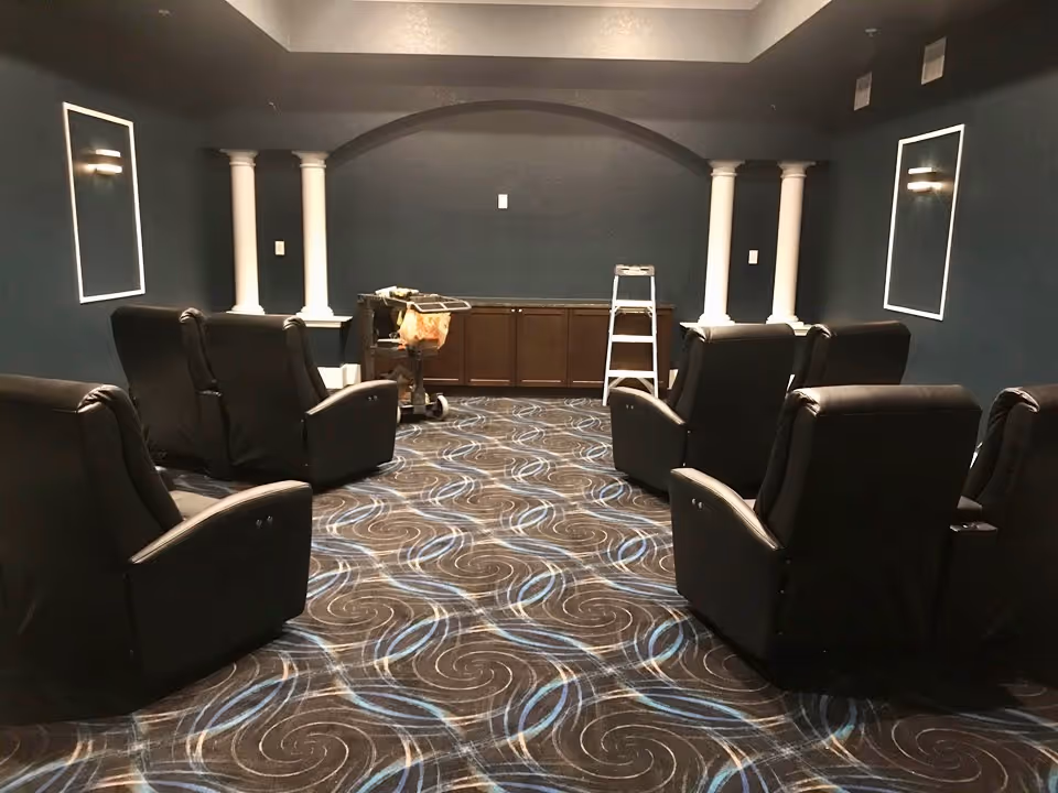 Small community theater-style room with black reclining chairs arranged facing a front wall with decorative columns and a ladder.