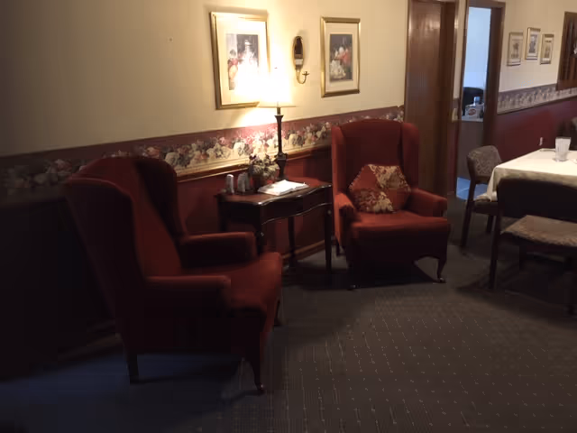 A cozy interior room with two red upholstered armchairs, a small wooden side table with a lamp and decorative items, floral wallpaper border, framed pictures on the wall, and a dining table with chairs in the background.