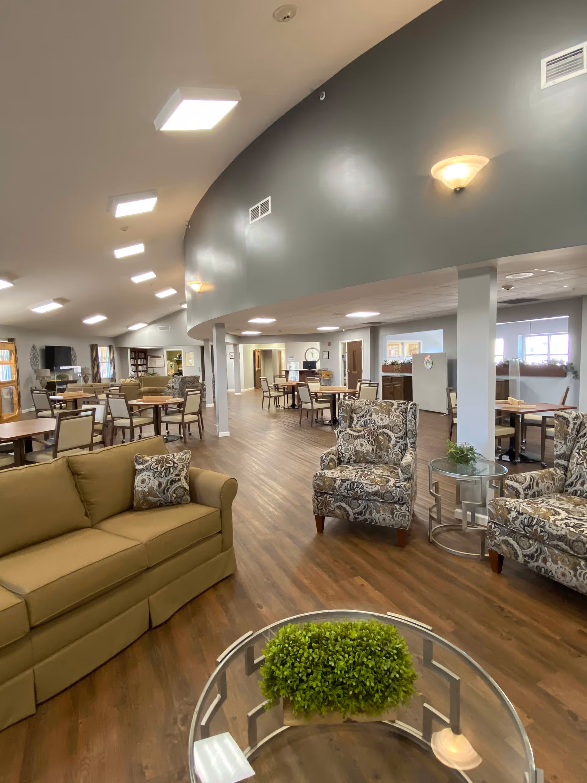 Spacious common area in an assisted living facility with wooden flooring, beige sofa, patterned armchairs, round glass coffee table with a green plant, and multiple dining tables and chairs in the background under bright ceiling lights.