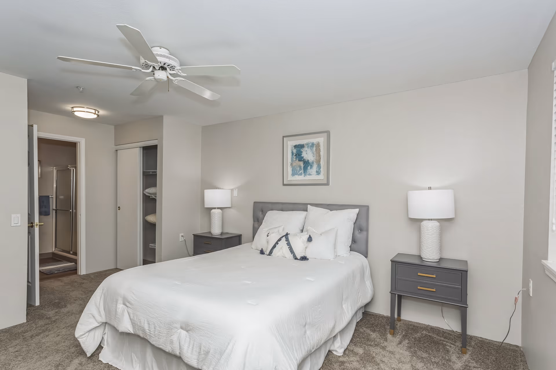 Bright neutral bedroom with a bed, two bedside tables and lamps, a ceiling fan, and an open closet.