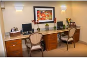 A well-lit office or work area with a long wooden desk featuring two computer monitors, two chairs with cushioned seats, a basket with papers, decorative plants, and a framed floral painting on the wall above the desk.