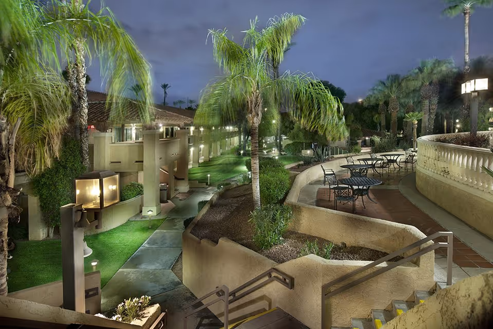 Landscaped outdoor courtyard at night with palm trees, illuminated walkways, stairs and patio tables.