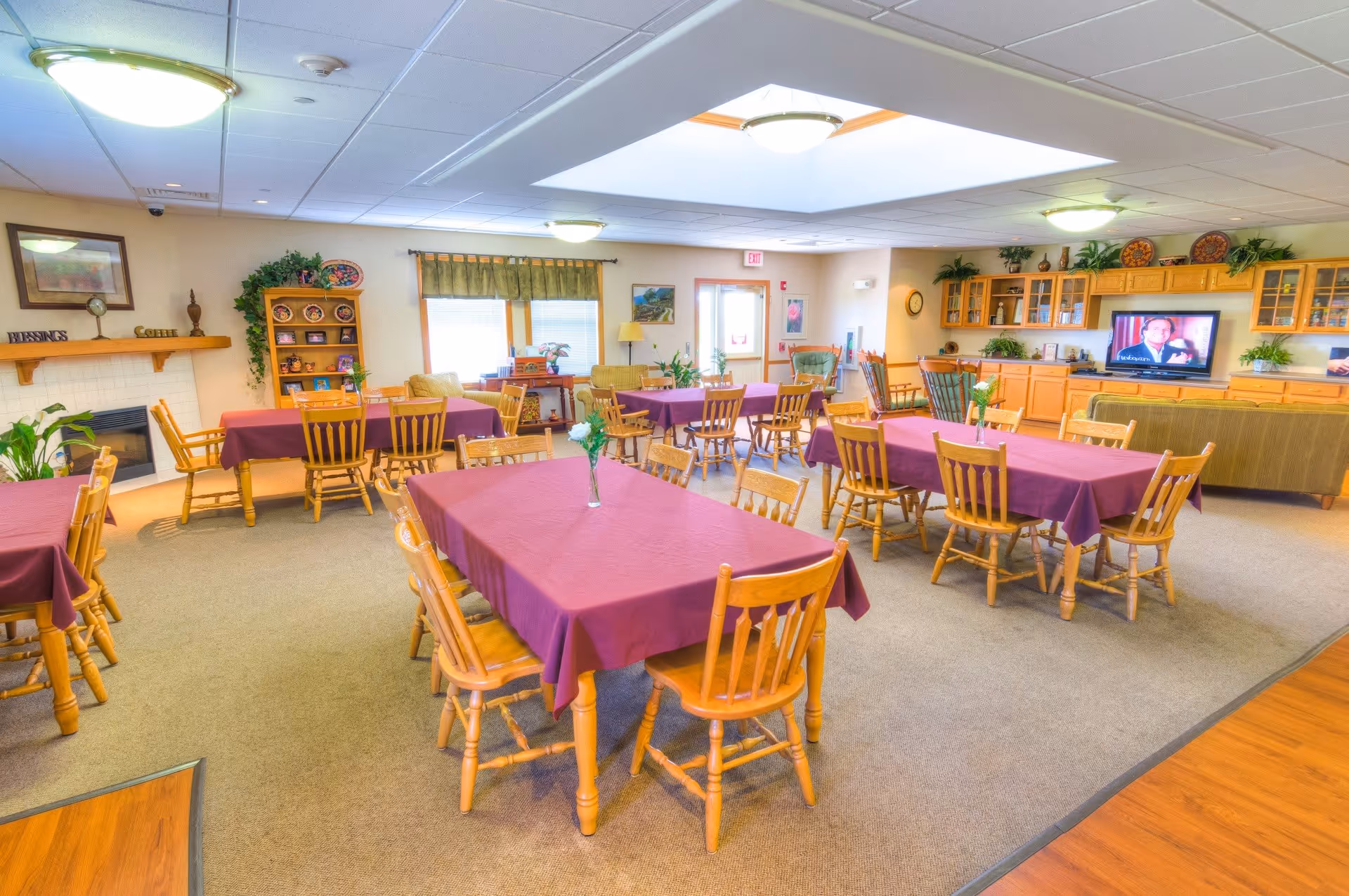 A bright and spacious dining room with multiple wooden tables covered with purple tablecloths, each surrounded by wooden chairs. The room features a fireplace on the left wall, a wooden cabinet with decorative plates, large windows with green curtains, and a television on the right side above a wooden cabinet. The ceiling has several round light fixtures, and there are plants and decorative items throughout the room.
