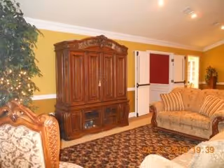 A cozy living room with yellow walls, a large wooden entertainment cabinet, a patterned area rug, and upholstered sofas and chairs with floral and striped designs. There is a potted plant with string lights on the left side and a door with a red bulletin board on the wall.