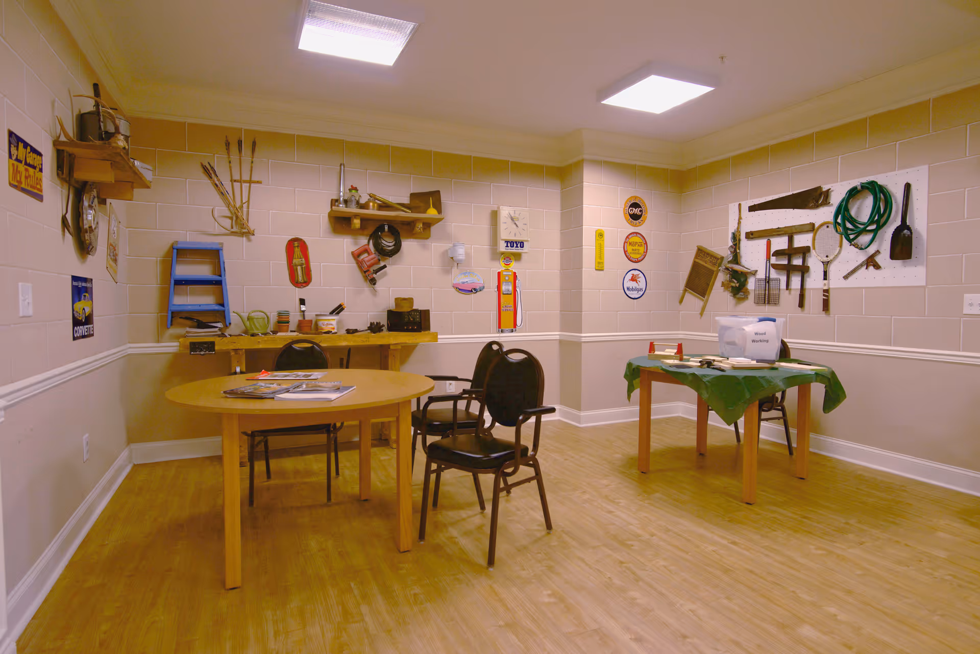 A room with light-colored walls and wooden flooring featuring two tables with chairs around them. One table has magazines on it, and the other is covered with a green cloth and has a white container labeled 'Wash Working.' Various vintage tools and signs are mounted on the walls, including a saw, a hose, and old-fashioned signs related to cars and gasoline.