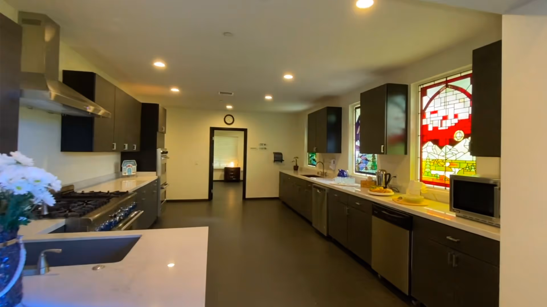 Long modern kitchen with dark cabinets, stainless steel appliances, white countertops and decorative stained-glass windows.