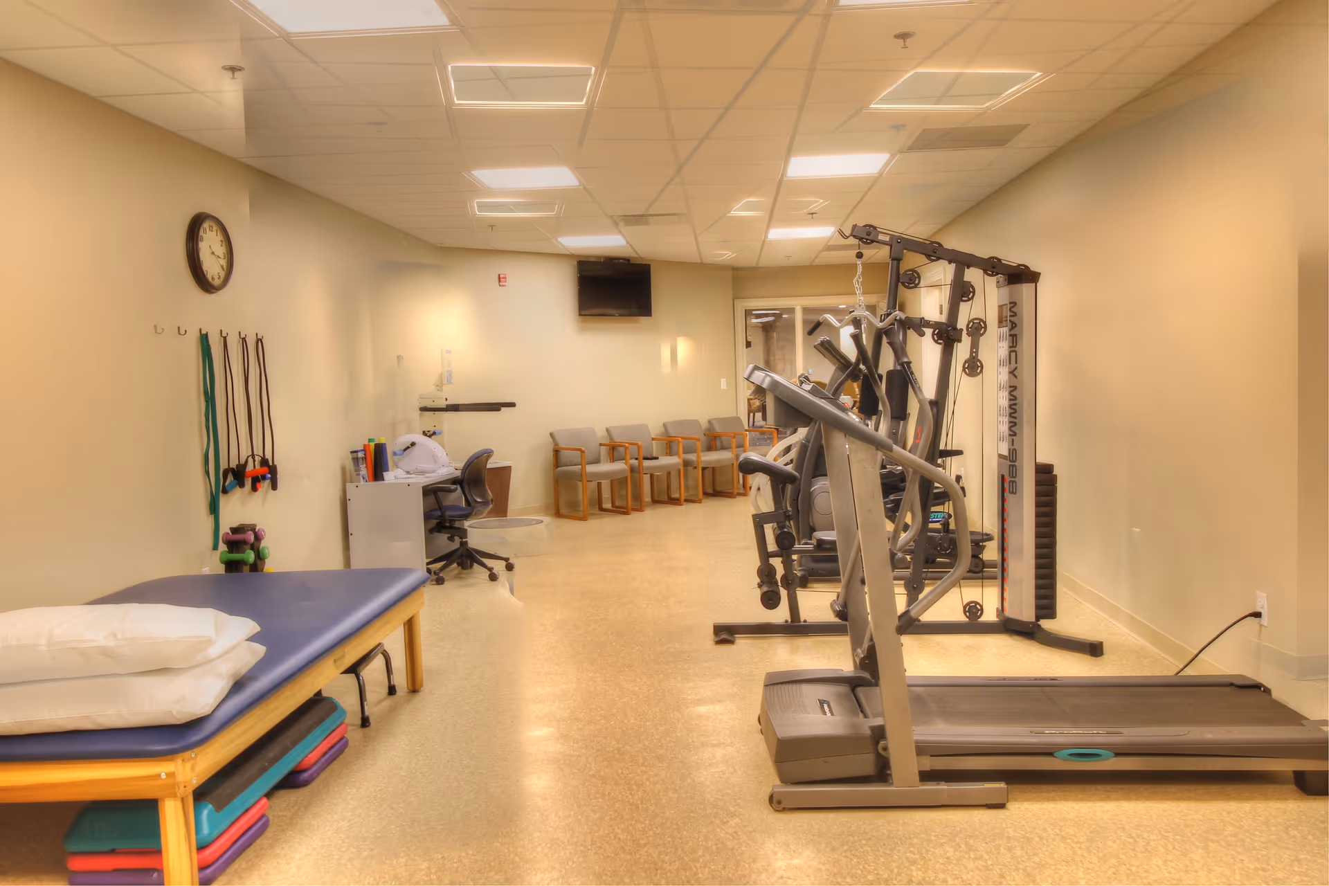 A well-lit rehabilitation and exercise room with treadmills, weight machines, a therapy table, resistance bands, and chairs.