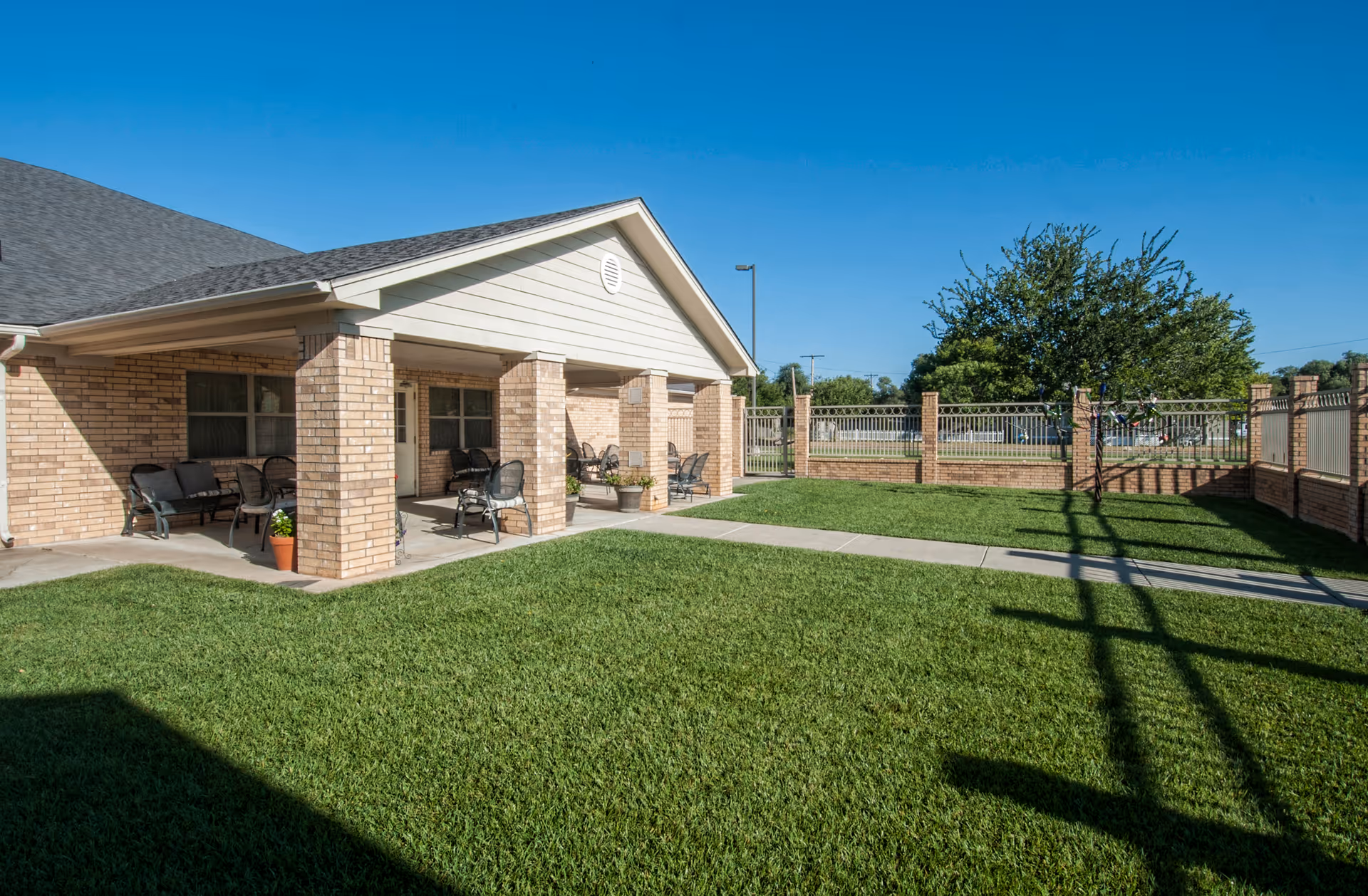Outdoor patio area of a brick building with a covered porch supported by brick columns. Several chairs and small tables are arranged under the porch. The area is surrounded by a green lawn and a brick and metal fence. A tree is visible near the fence under a clear blue sky.