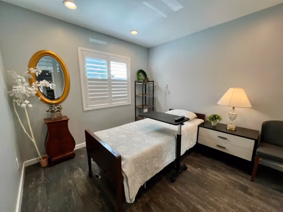 A small, neatly arranged bedroom with a single bed covered in a white quilt. The bed has an overbed table with a remote control on it. To the right of the bed is a nightstand with a lamp and a small potted plant. There is a gray chair next to the nightstand. On the left side of the room, there is a wooden chest with a decorative vase and a tall artificial white plant next to it. Above the chest is a round gold-framed mirror. The room has gray walls, dark wood flooring, and a window with white shutters letting in natural light.