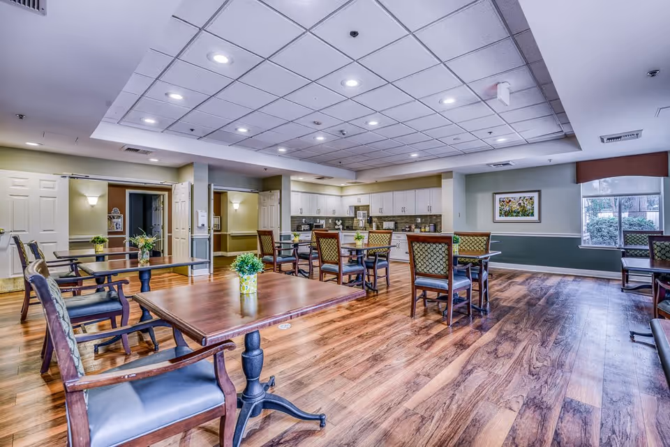 A spacious dining room with wooden floors, multiple tables and chairs arranged neatly, small potted plants on each table, white cabinets and countertops along the back wall, a window with a view of greenery, and soft ceiling lighting.