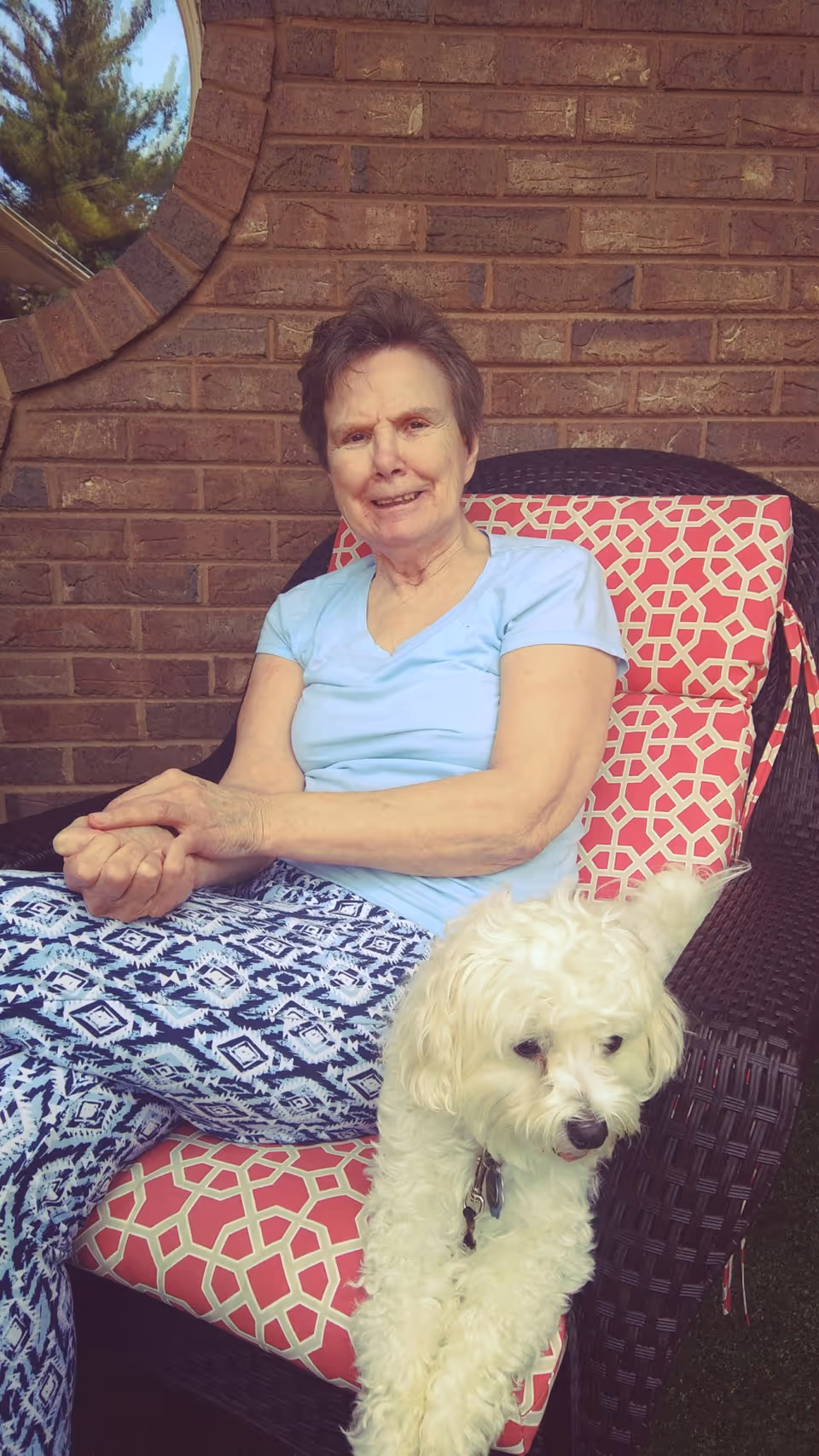 An elderly woman sitting on a cushioned outdoor chair with a red and white geometric pattern, smiling gently. A small white fluffy dog is resting on the arm of the chair. The background shows a brick wall and part of a circular window with trees visible outside.