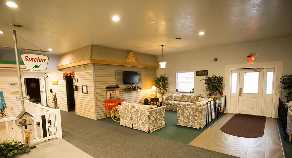 Interior view of a senior living facility common area with floral patterned couches arranged around a small table, a popcorn machine, a mounted TV, and decorative plants near a set of double doors with exit signs.