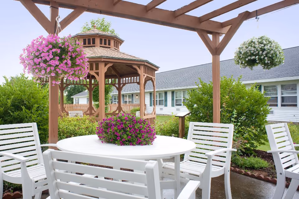 Outdoor seating area with white chairs and a round table under a wooden pergola. Hanging flower baskets with pink and white flowers are attached to the pergola. In the background, there is a wooden gazebo surrounded by greenery and a single-story building with multiple windows.