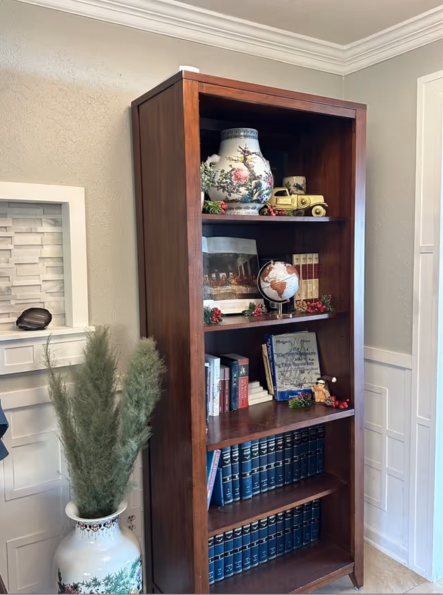 Tall wooden bookshelf in a living room corner holding decorative vases, a globe, books, and small ornaments with a large floral vase of greenery beside it.