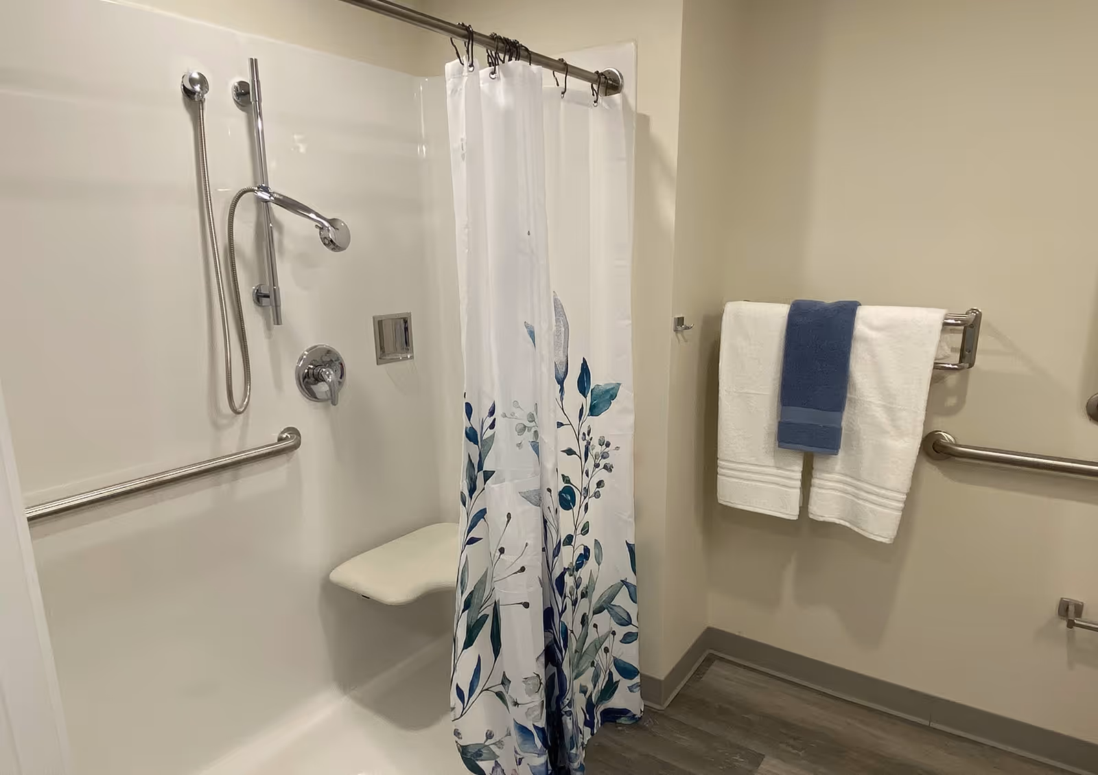 A bathroom shower area with a white wall, a mounted showerhead, a handheld shower wand, and a fold-down seat. There is a white shower curtain with blue and green leaf patterns partially drawn. On the adjacent wall, there are two white towels and one blue towel hanging on a towel rack, with grab bars installed for support.
