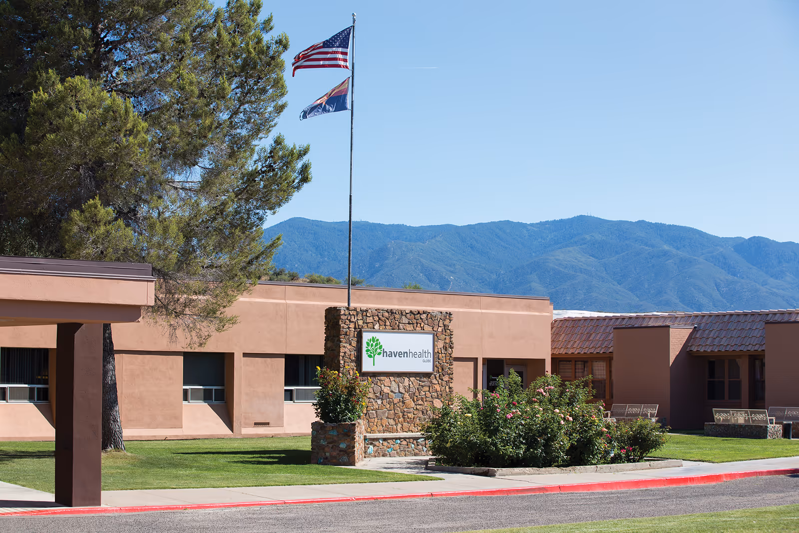 Exterior view of Haven Health Globe facility with a stone sign displaying the facility name, two flagpoles with American and state flags, surrounded by green grass, bushes, and trees, with mountains in the background under a clear blue sky.