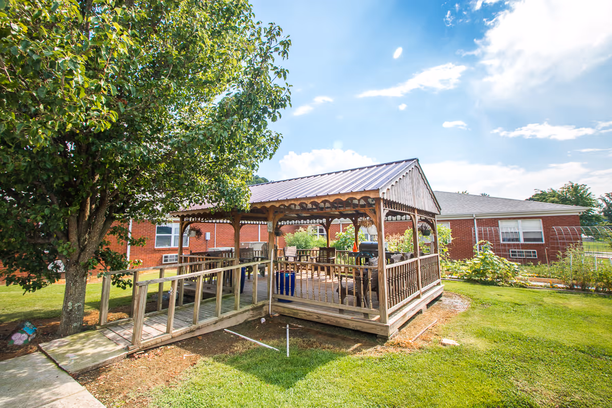 Outdoor wooden gazebo with a metal roof in a grassy yard next to a tree, featuring chairs and tables inside. A red brick building is visible in the background under a partly cloudy sky.