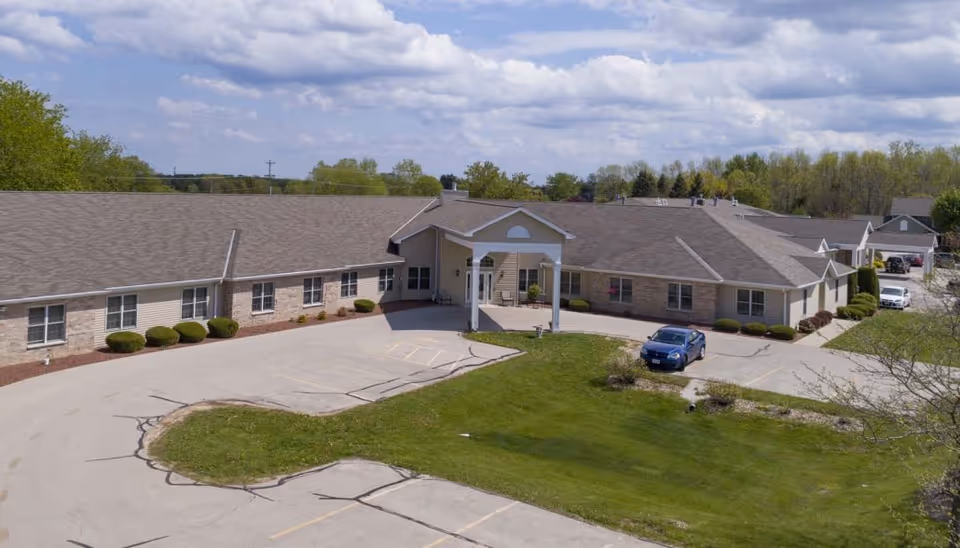 Exterior view of a single-story assisted living facility building with a covered entrance, surrounded by a parking lot and green lawn under a partly cloudy sky.