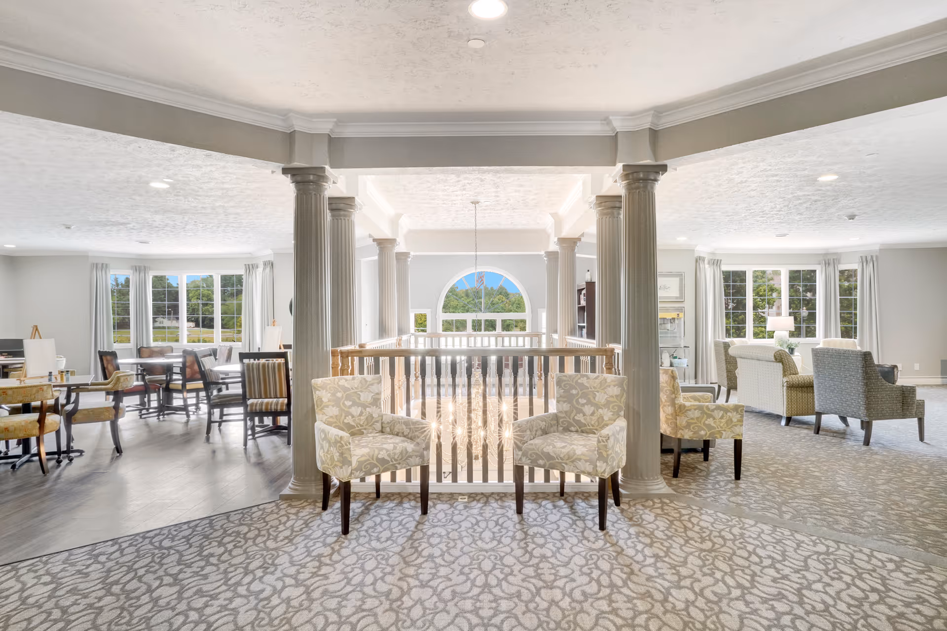 Bright and spacious common area in a senior living facility with patterned carpet, multiple seating arrangements including floral armchairs and sofas, large windows letting in natural light, and decorative columns framing a railing overlooking a lower level with a chandelier.