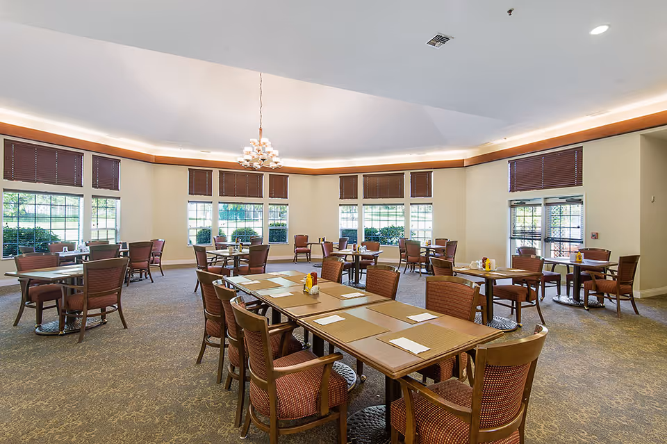 A spacious dining room with multiple tables and chairs arranged neatly. The room has large windows with brown blinds allowing natural light to enter. The tables are set with placemats, napkins, and condiments. A chandelier hangs from the ceiling, and the carpet has a subtle pattern.