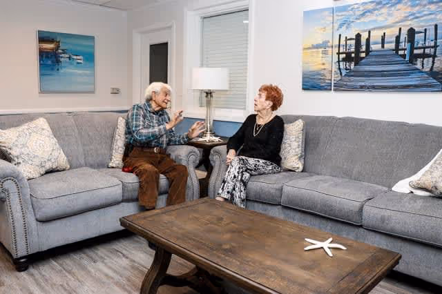 Two elderly people sitting on gray couches in a living room, engaged in conversation. The room features a wooden coffee table with a starfish decoration, a side table with a lamp, and two framed pictures of docks over water on the walls.