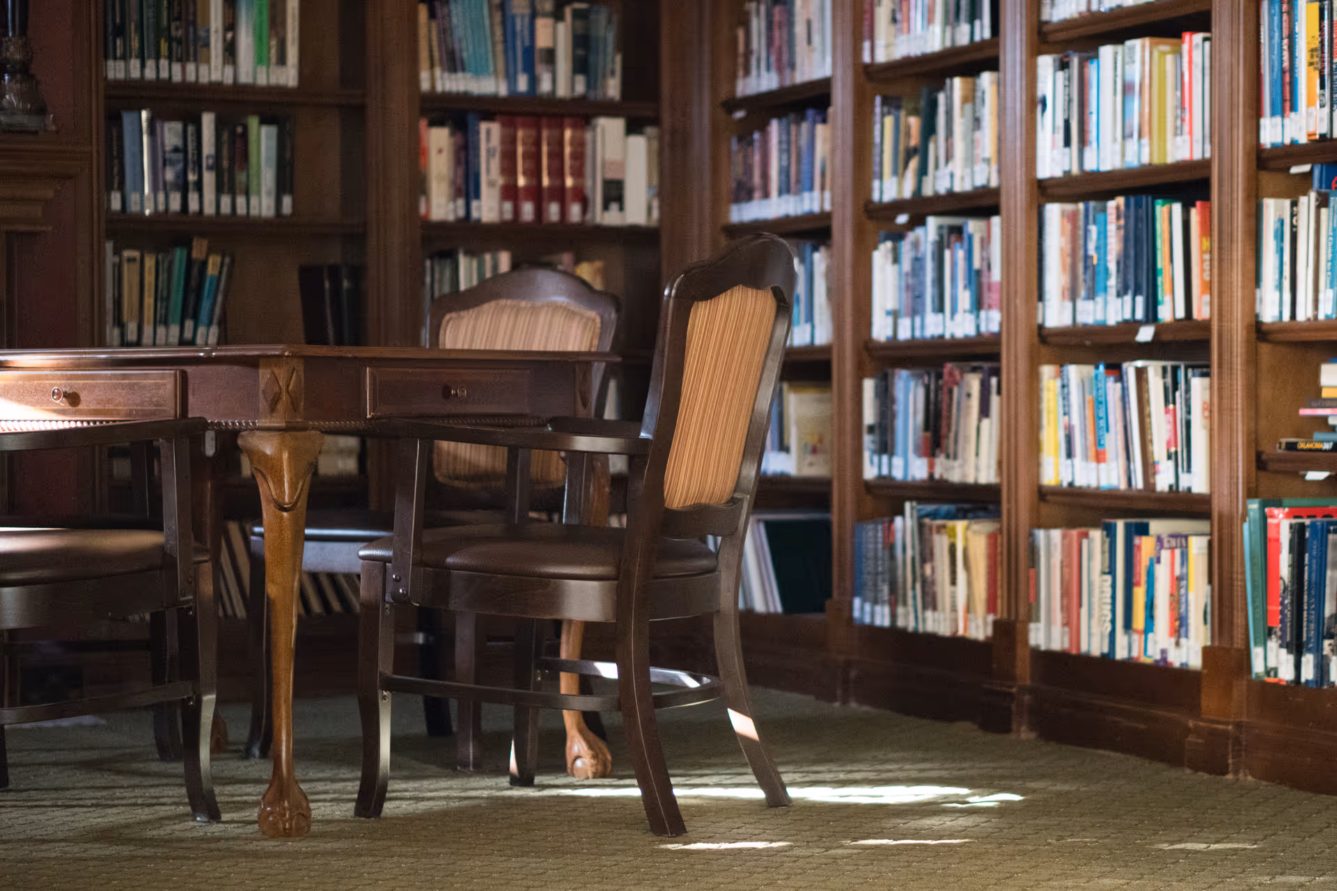 A quiet library room with wooden bookshelves filled with books lining the walls. In the center, there is a wooden table with carved legs and several wooden chairs with cushioned seats and backs arranged around it. The room has a warm, classic ambiance with soft lighting.