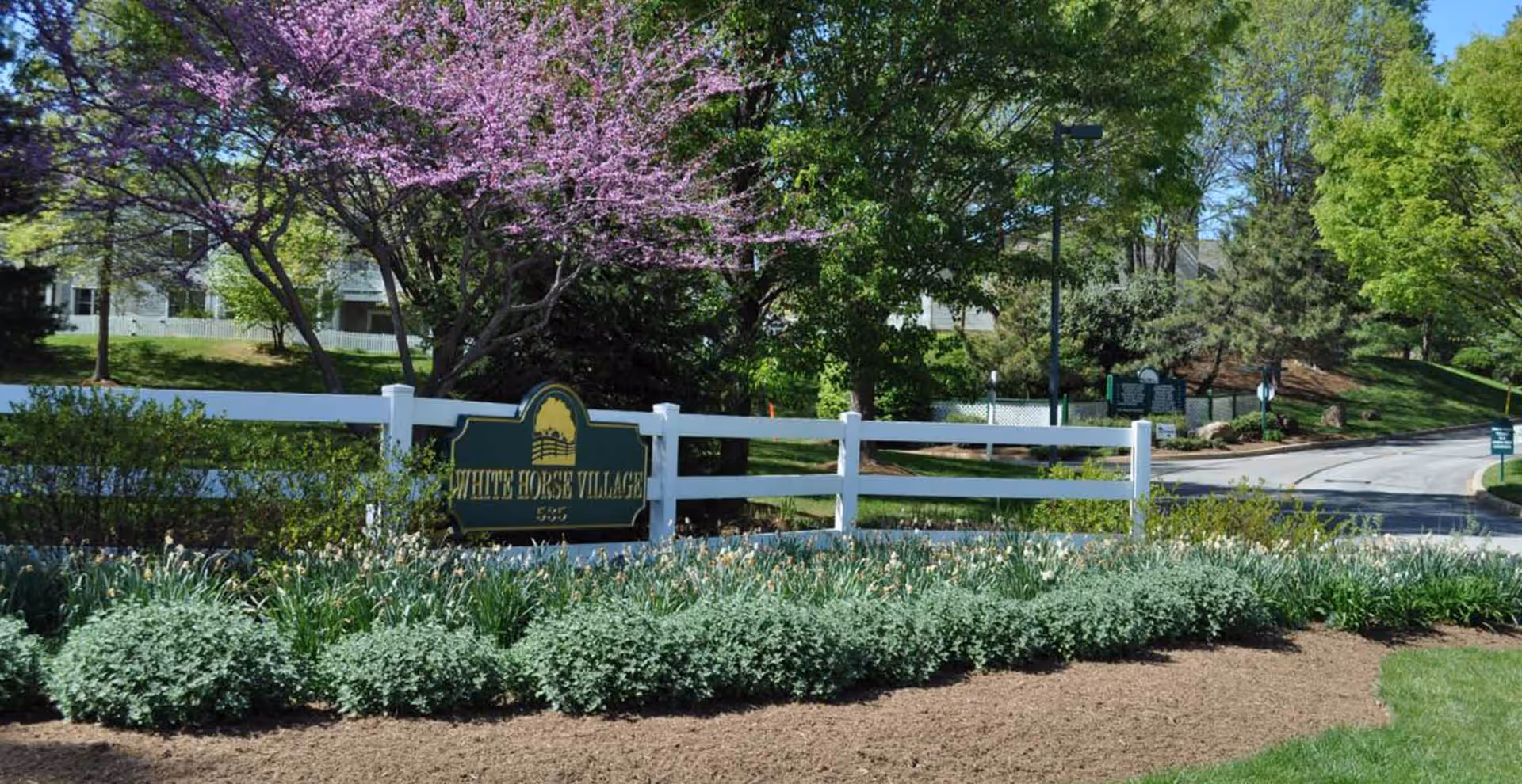 Entrance to White Horse Village featuring a white wooden fence with a green and yellow sign displaying the facility name. Surrounding the sign are well-maintained bushes and flowering plants, with trees in full bloom including one with pink flowers. A paved road curves to the right in the background under a clear blue sky.