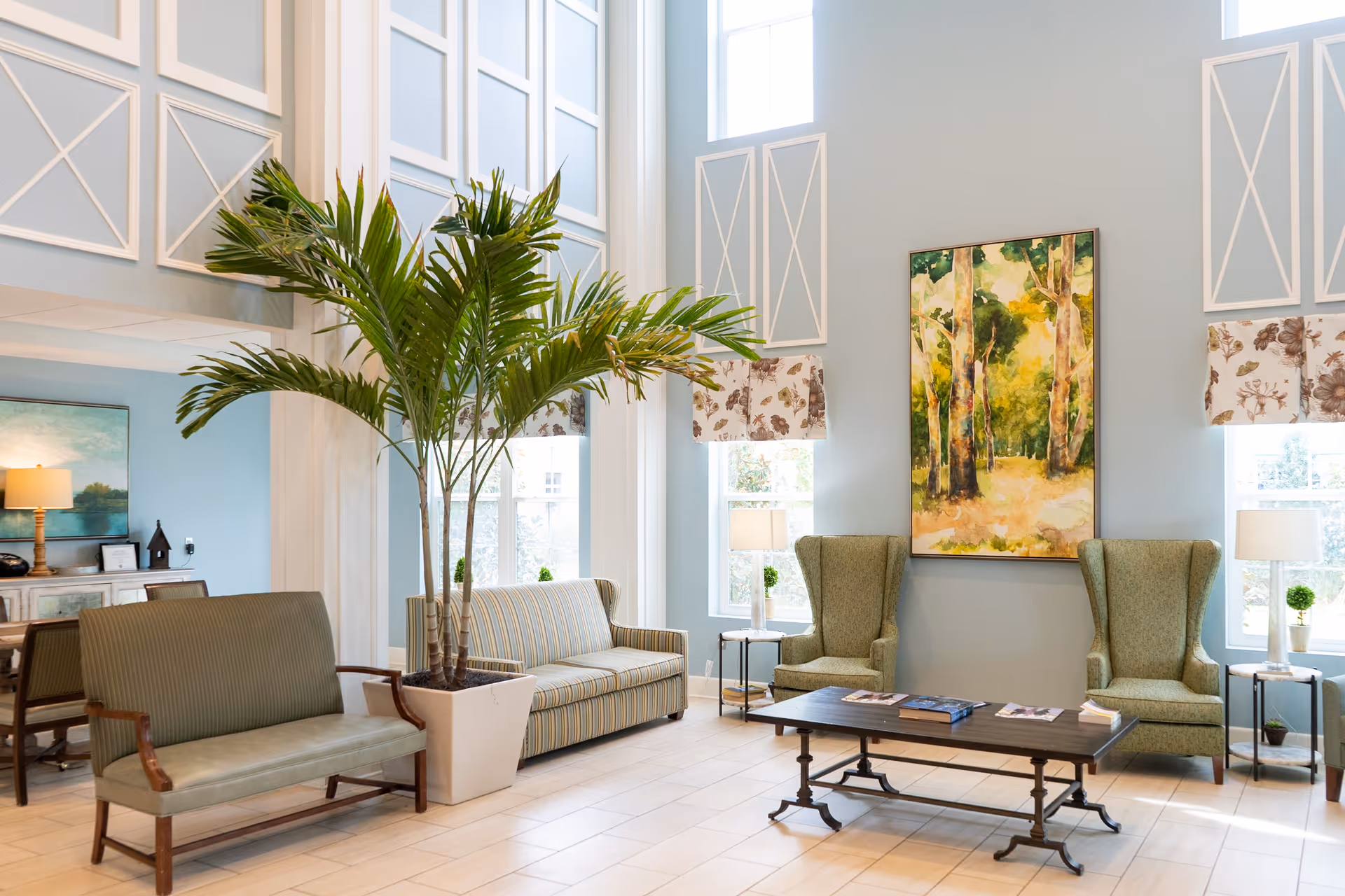 A bright and spacious living room area with high ceilings and large windows. The room features two green upholstered armchairs, a striped sofa, a green bench, a large potted palm plant, a wooden coffee table with magazines, and a colorful painting of a forest scene on the wall. There are also two side tables with lamps and small plants, and floral valances on the windows.