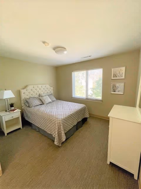 A bedroom with a neatly made bed featuring a tufted headboard and patterned bedding. There is a white nightstand with a lamp and some items on it to the left of the bed. A window with blinds lets in natural light, and two framed pictures hang on the wall to the right of the window. A white dresser is positioned against the right wall.