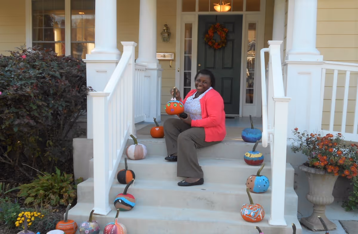 A woman sitting on the front steps of a house decorated with various painted pumpkins. The house has white railings, columns, and a dark green door with a fall wreath. There are bushes and flowers on either side of the steps.