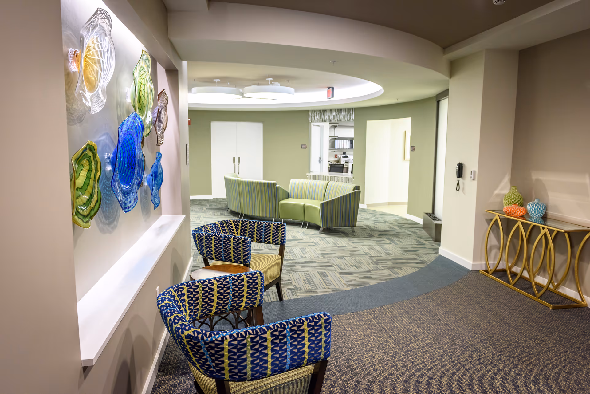 Interior view of a senior living facility lounge area with patterned blue and green chairs arranged along a curved wall. The wall features colorful glass art pieces. In the background, there are green and striped sofas under a ceiling with circular light fixtures. A decorative gold console table with colorful vases is on the right side.