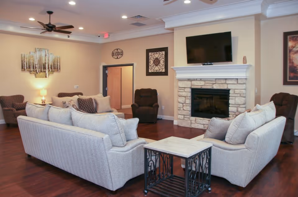 A cozy living room with two light gray sofas arranged around a small square table with a metal frame. The room features a stone fireplace with a flat-screen TV mounted above it. There are two brown armchairs, a ceiling fan, and wall decorations including a painting of trees and a decorative metal piece. The floor is wooden, and the walls are painted beige.