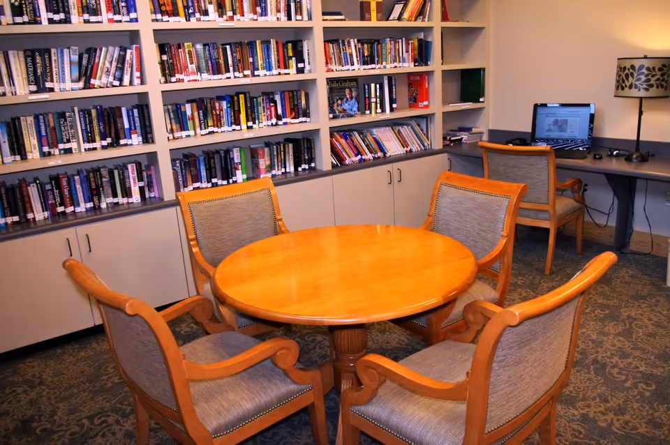 A cozy library or reading room with a round wooden table surrounded by four cushioned wooden chairs. Behind the table, there are shelves filled with books and cabinets below. To the right, there is a desk with a computer and a decorative lamp on it.