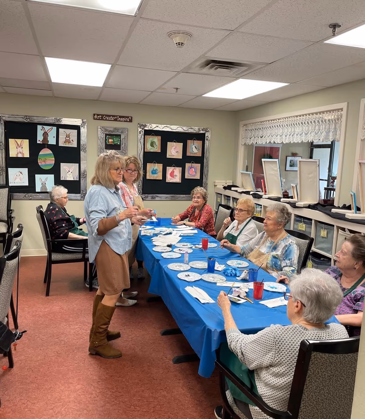 A group of elderly women seated around a table covered with a blue tablecloth, participating in an art activity with paintbrushes and paper plates. Two women stand at the side of the table, one speaking to the group. The room has framed artwork on the walls and a window with lace curtains.