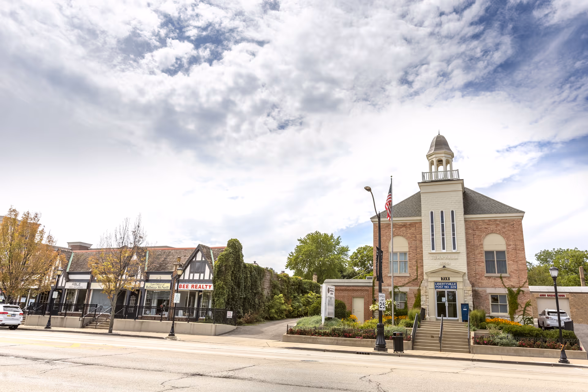 Street view of a small commercial area featuring a Tudor-style building with signs for Bee Realty and other businesses on the left, and a brick building with a cupola and an American flag on the right, under a partly cloudy sky.