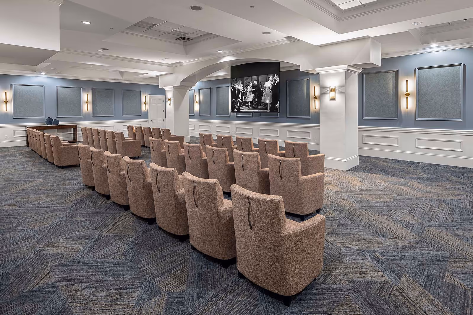 A spacious room with rows of beige upholstered chairs facing a large wall-mounted television screen displaying a black and white image. The room features blue walls with decorative panels, modern wall sconces, and a patterned carpet floor. The ceiling has recessed lighting and architectural details.