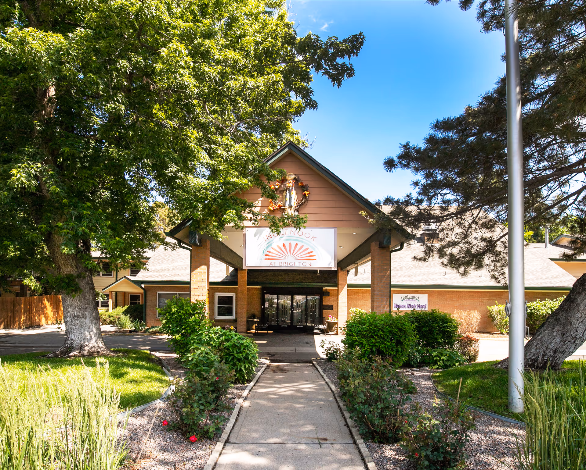 Front entrance of Inglenook At Brighton facility with a covered walkway, surrounded by green trees and bushes under a clear blue sky.